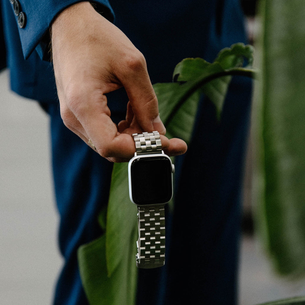 Hand in blue suit holding a silver smartwatch with a metal link band above green plant leaves.