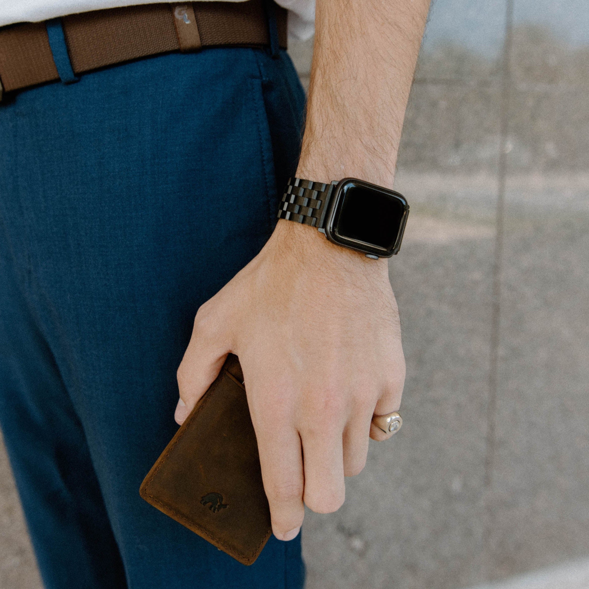 Man's wrist wearing black metal link Apple Watch strap, hand holding brown leather card wallet