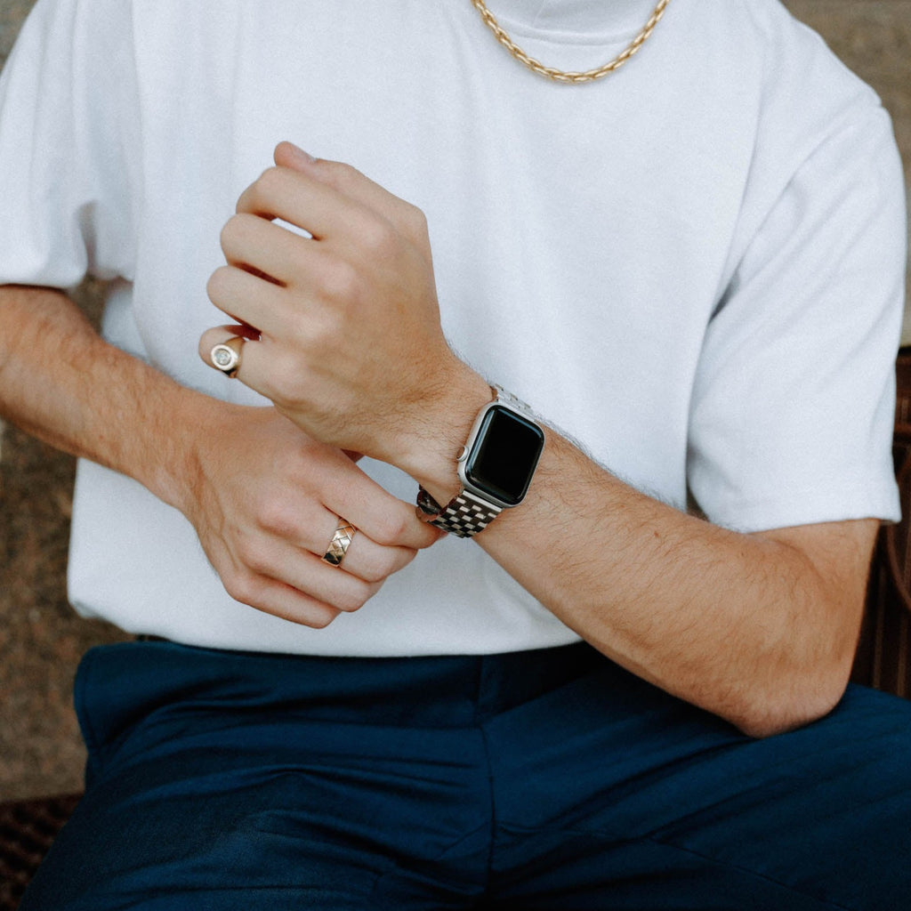 Male torso and hands wearing a silver smartwatch, two gold rings and a chain; white tee, navy pants, brown leather bag