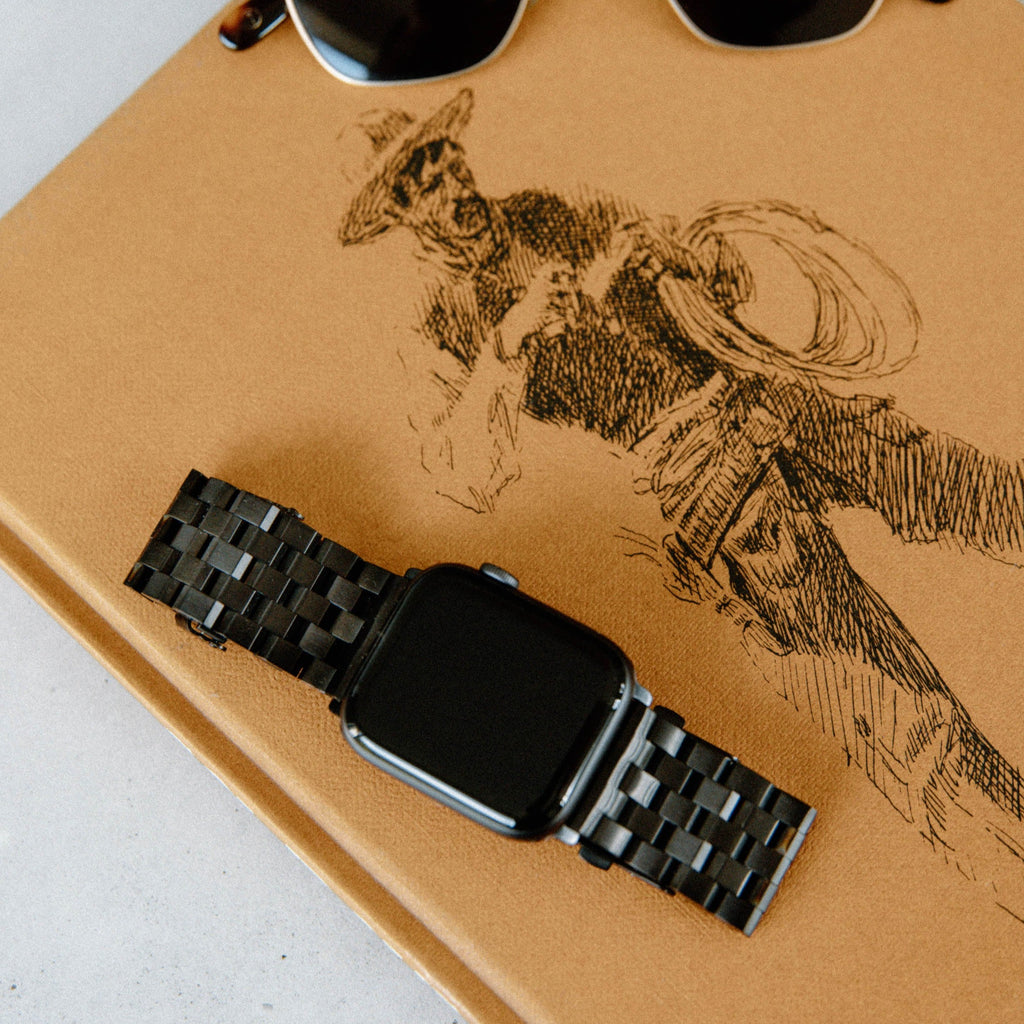 Black stainless steel link Apple Watch band laid across tan leather notebook with a cowboy sketch