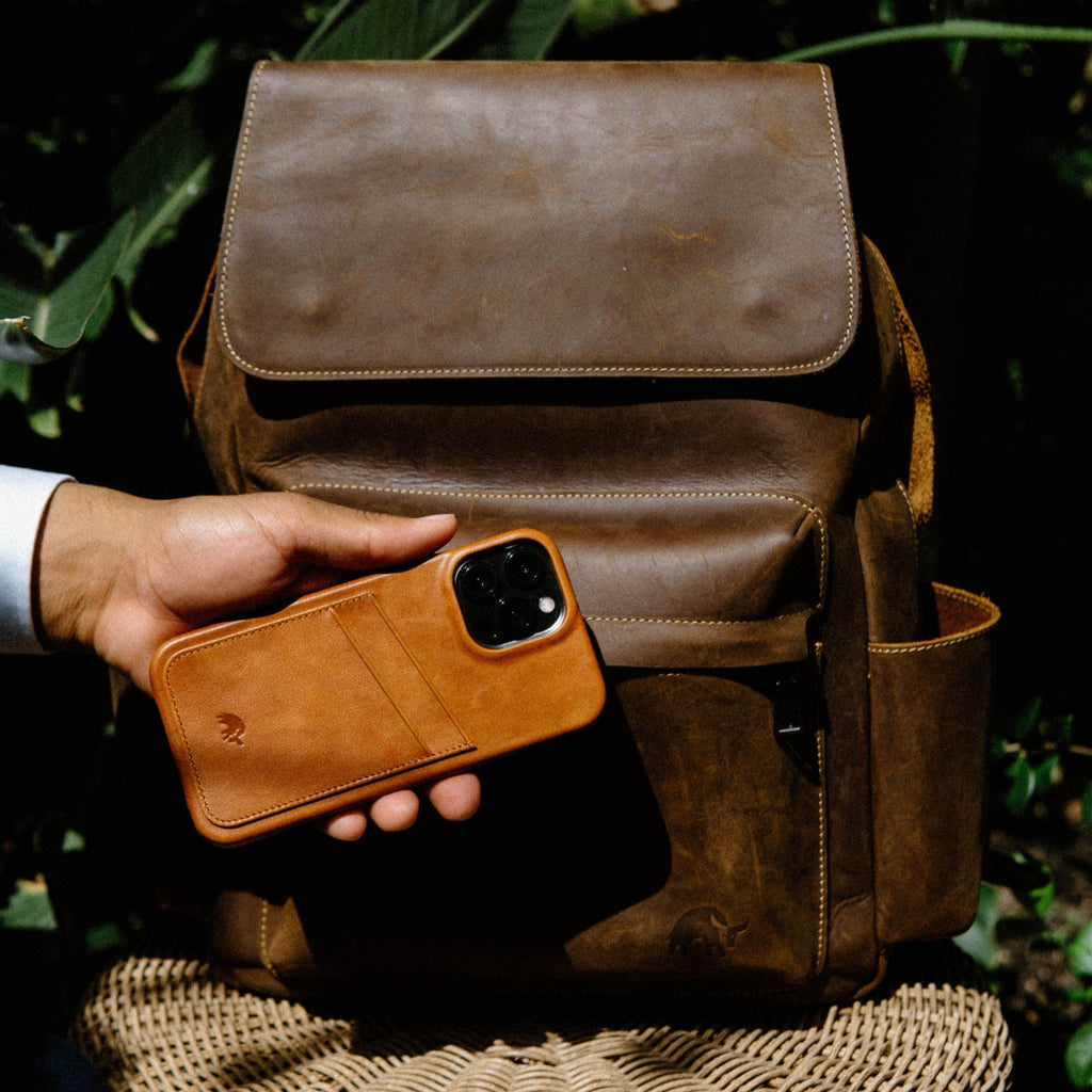 Hand holding tan leather card-wallet iPhone case with card slot in front of brown leather backpack