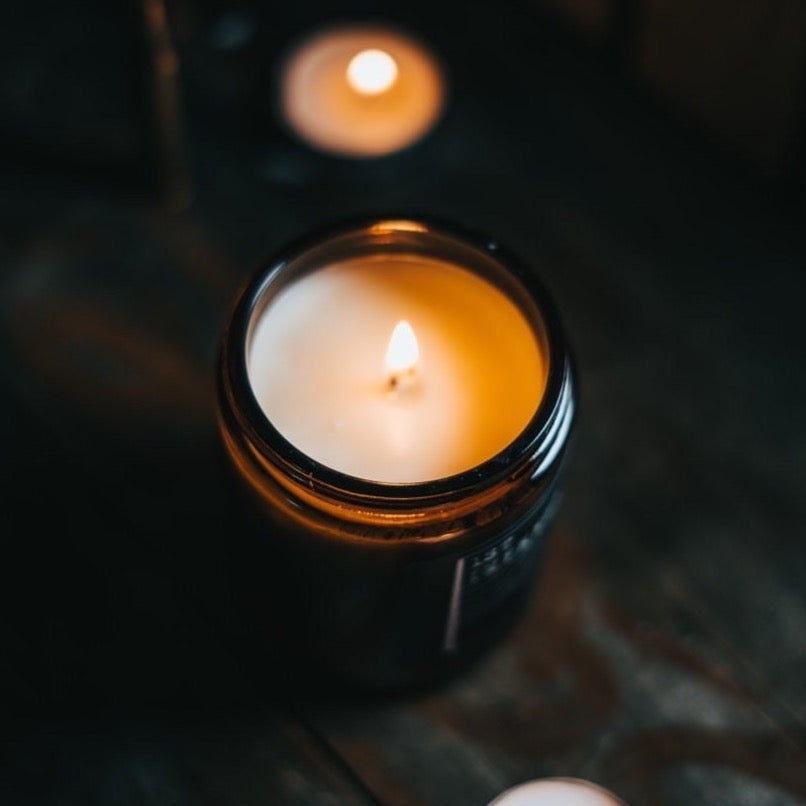 Lit candle burning in a brown glass jar, casting a warm amber glow in a dark setting.